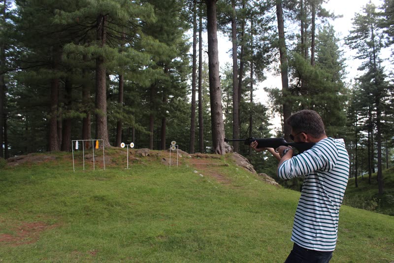 A visitor aiming an air rifle at targets on a grassy slope under the pines