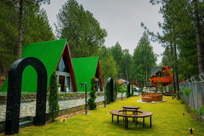 Green-roofed A-frame cabins around a lawn with picnic tables at TDCP Chewra Base Resort