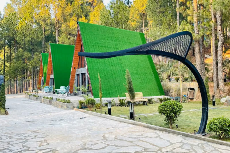 A row of green A-frame cabins along a paved courtyard with garden beds