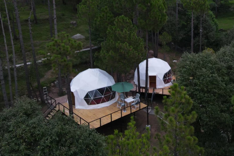 Aerial view of two white glamping domes on wooden decks among pine trees at Khanabadosh Glamps Murree